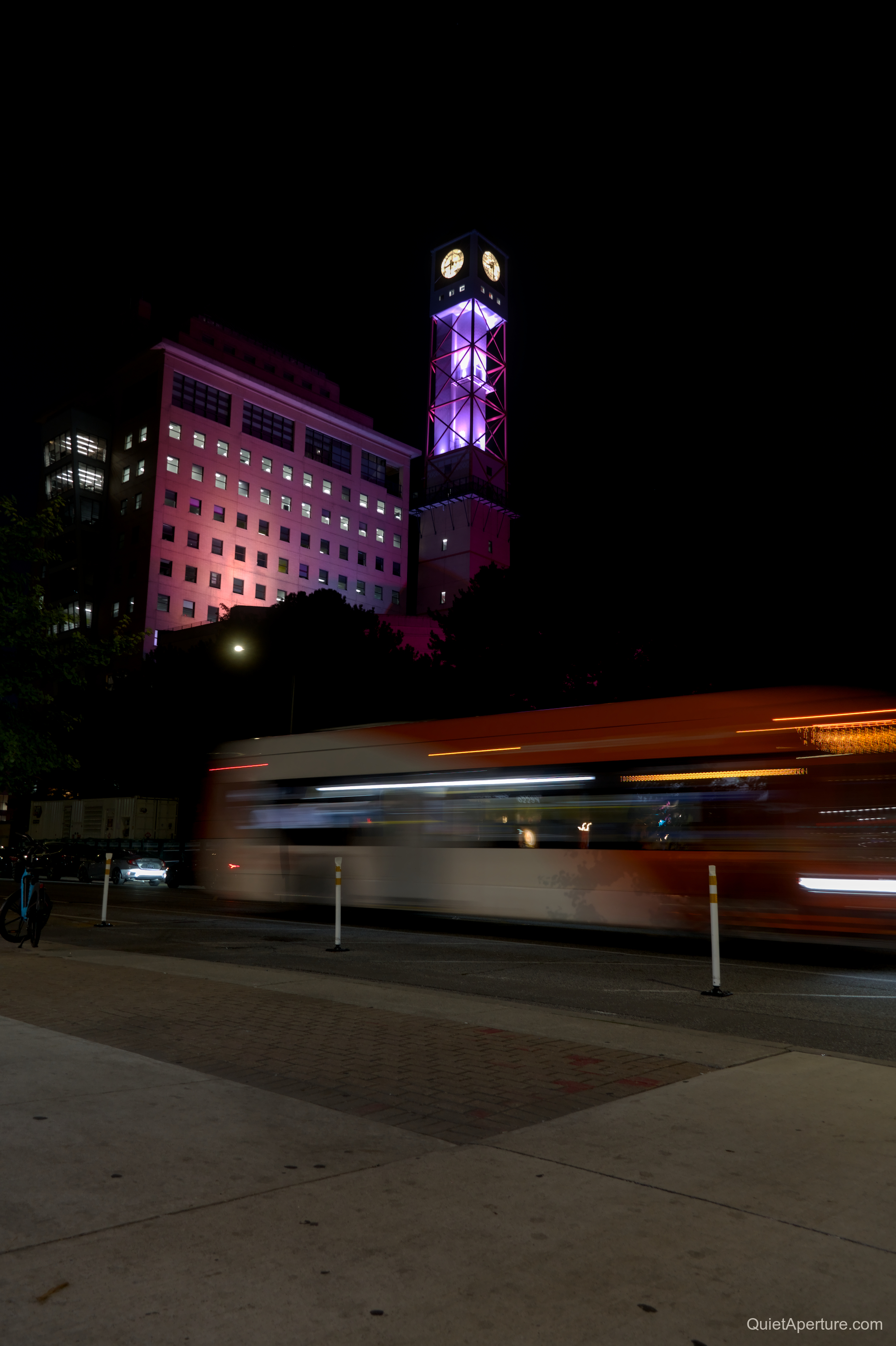 A passing bus beneath a purple-lit clock tower at night.